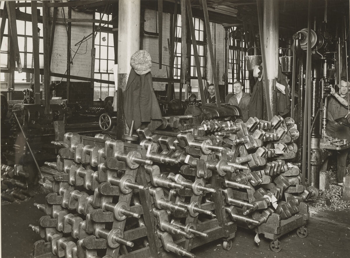 Oude zwart-witfoto van rekken vol krukassen in een fabriekshal, met arbeiders erachter