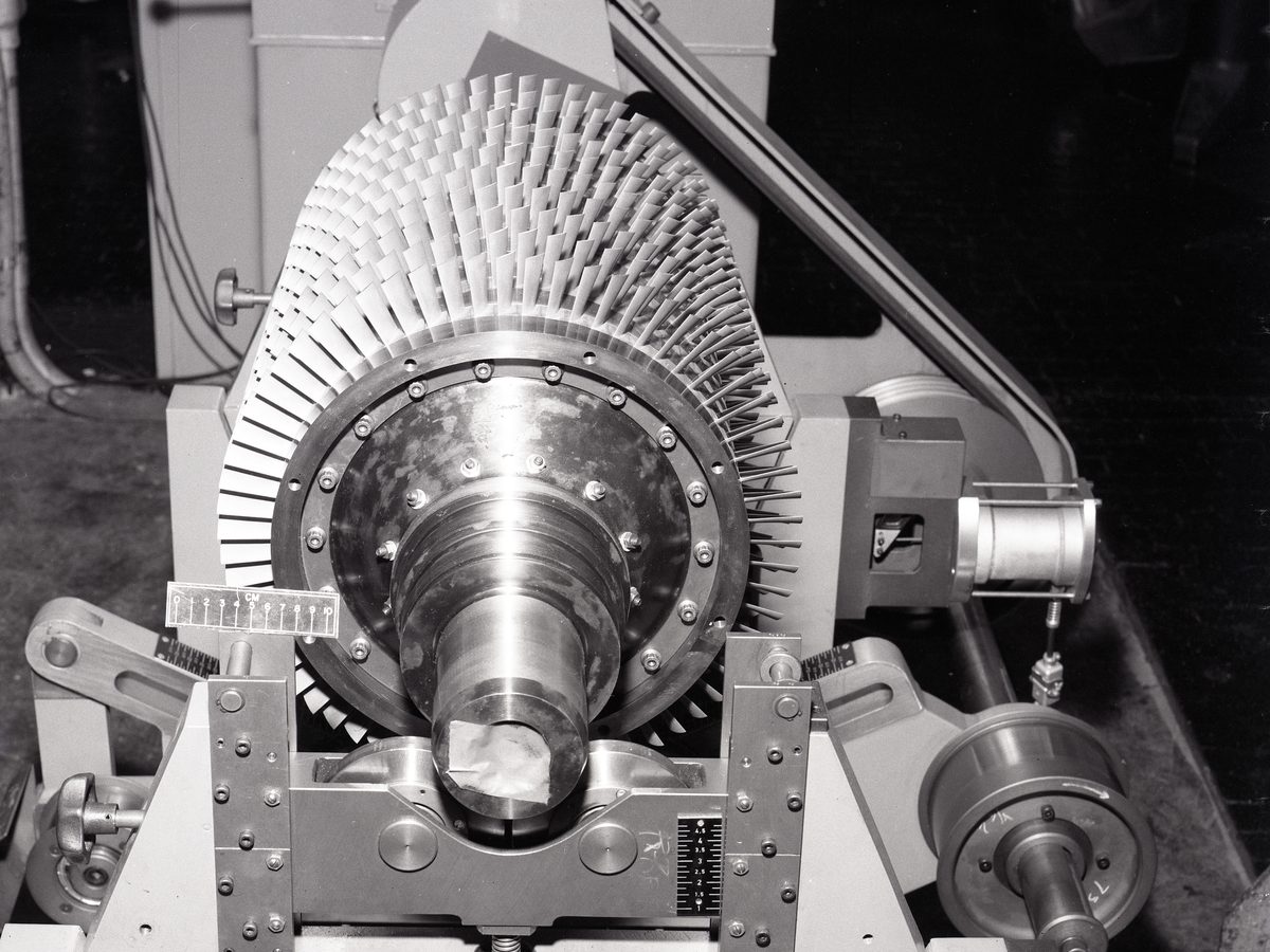 Een rotor met schoepen ligt in de lagerbokken van een balanceermachine, met de aandrijfriem erachter
