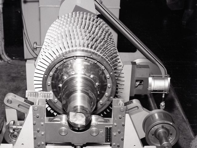 Een rotor met schoepen ligt in de lagerbokken van een balanceermachine, met de aandrijfriem erachter